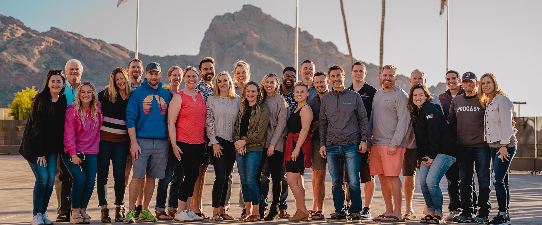A large group of smiling adults pose together outside near palm trees and flag poles, with dry mountains behind them.