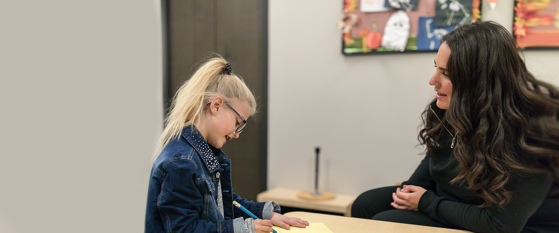 An elementary instructor sits at and leans over a table, conversing with a child who is working on handwriting with a large pencil.