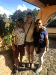 Three students stand close together, smiling, with greenery and a house in the background.