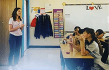 A teacher stands at the front of a classroom, speaking to students seated at desks, with 