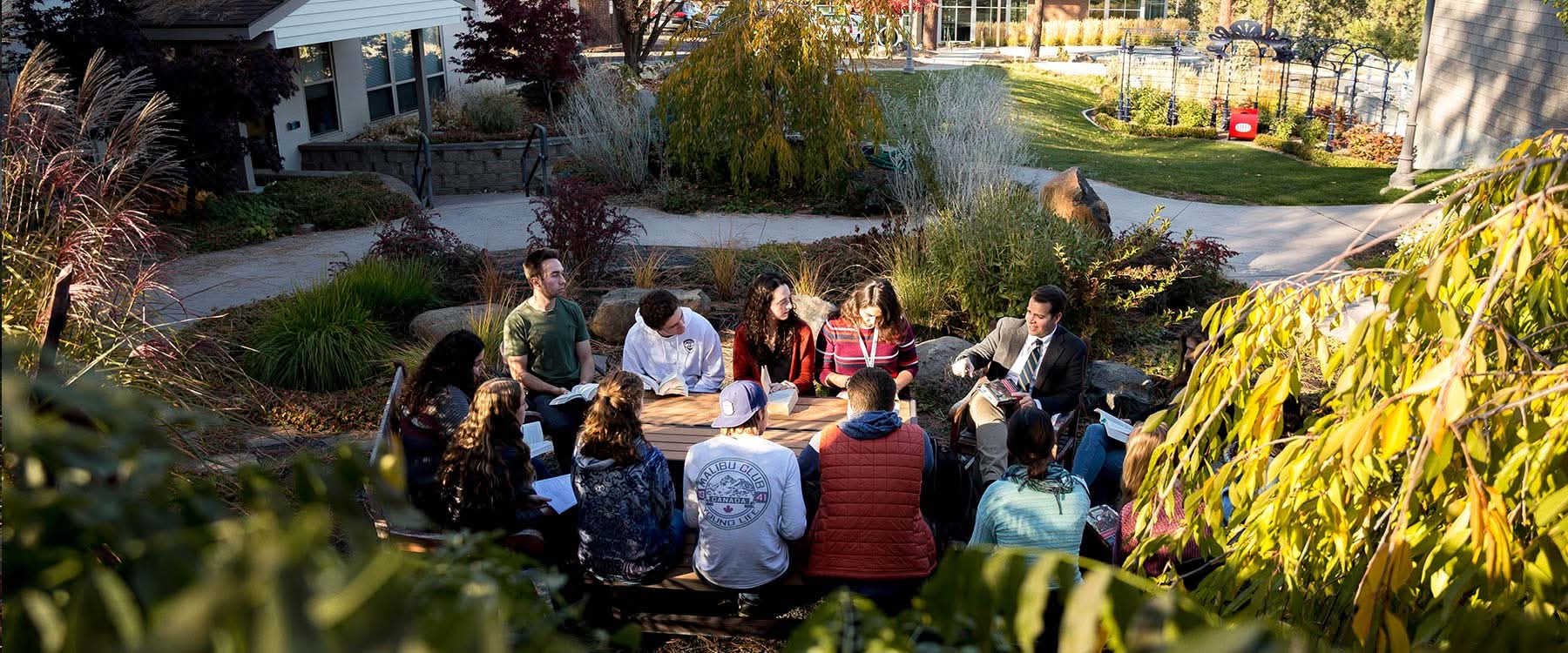 Multiple students sit around an outdoor table on campus, listening to an engaging professor.