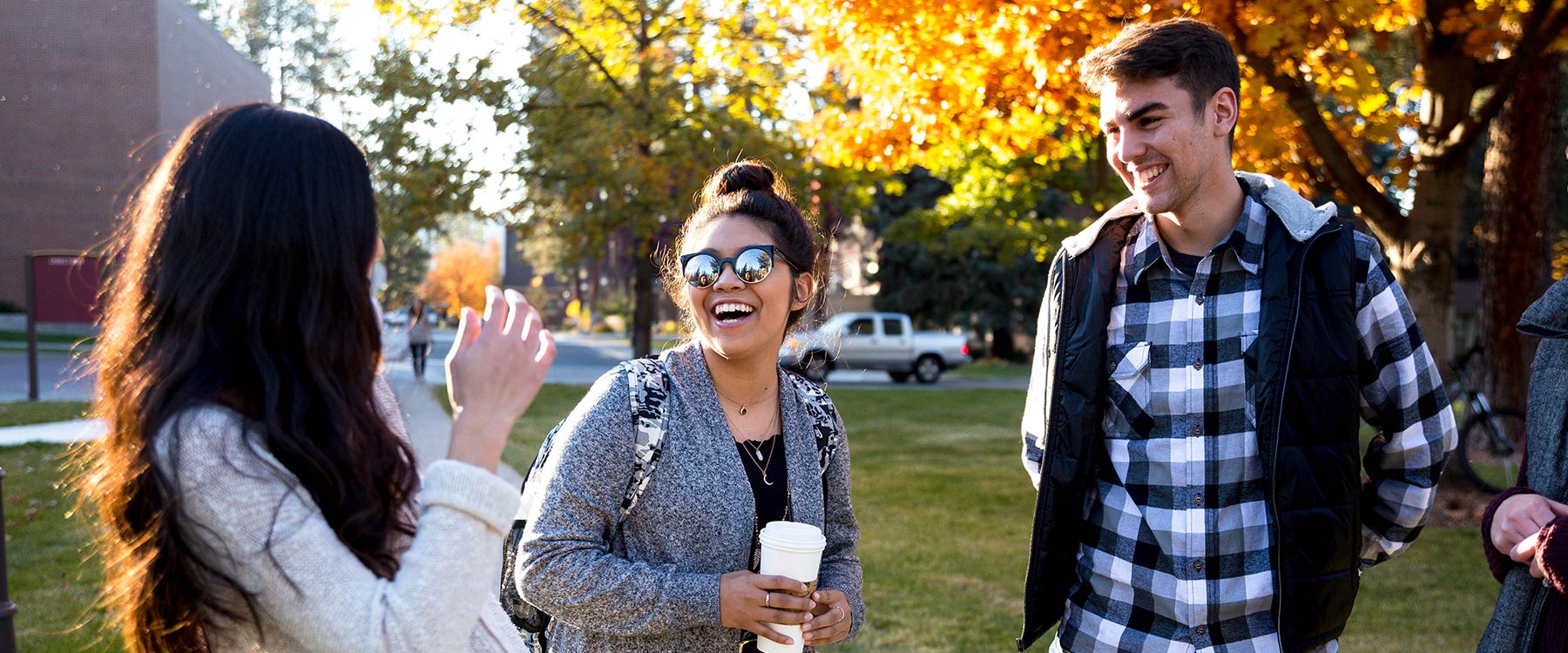 Three students stand outdoors on campus, chatting and smiling, on a sunny autumn day.