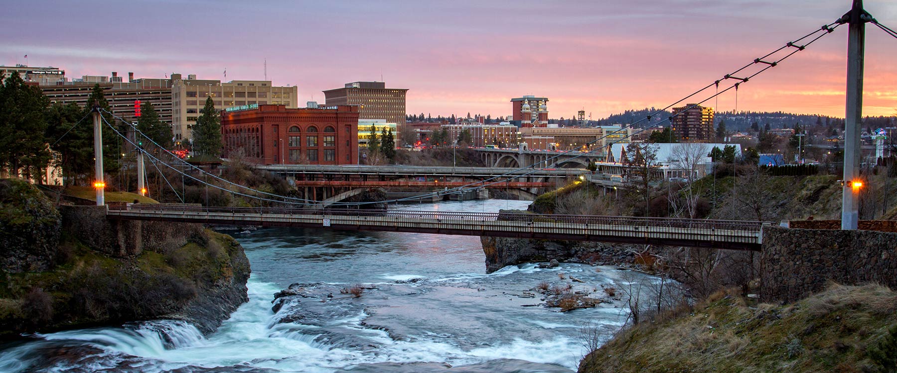 Spokane river flowing underneath a bridge, with the city skyline in the background at sunset.