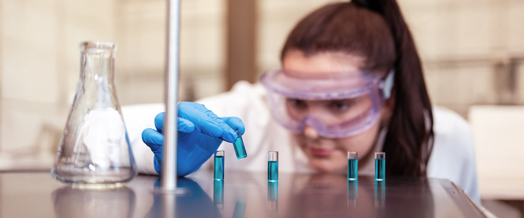 A science student in goggles reaches for vials with blue liquid in a lab setting.