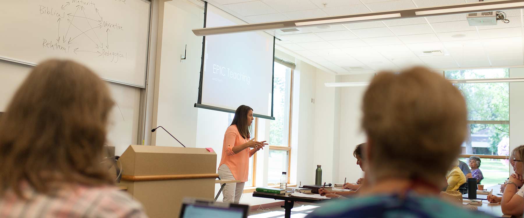 A professor stands at the front of a classroom and speaks to a full class of students.