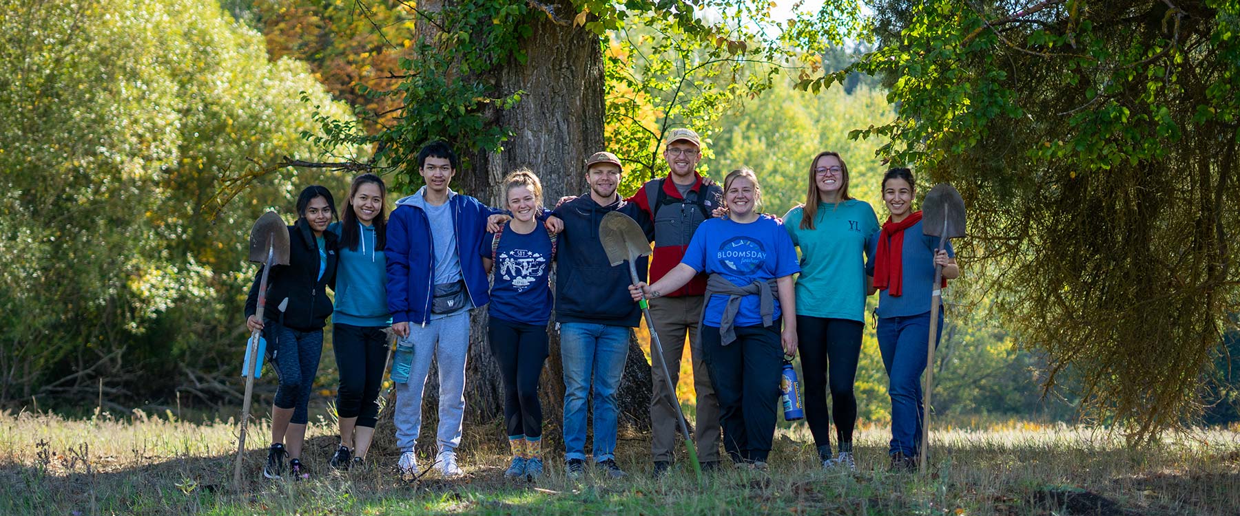 A group of student volunteers poses in front of a tree, holding shovels and smiling.
