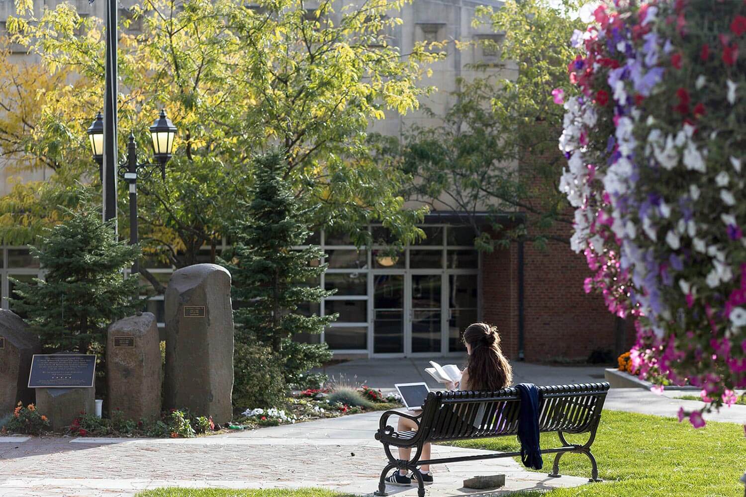 A student with long hair is seen from behind, sitting on a bench, studying with a laptop and book.