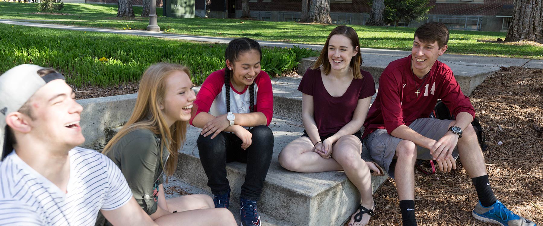 A group of students sit on a set of concrete steps on campus smiling, talking and laughing.