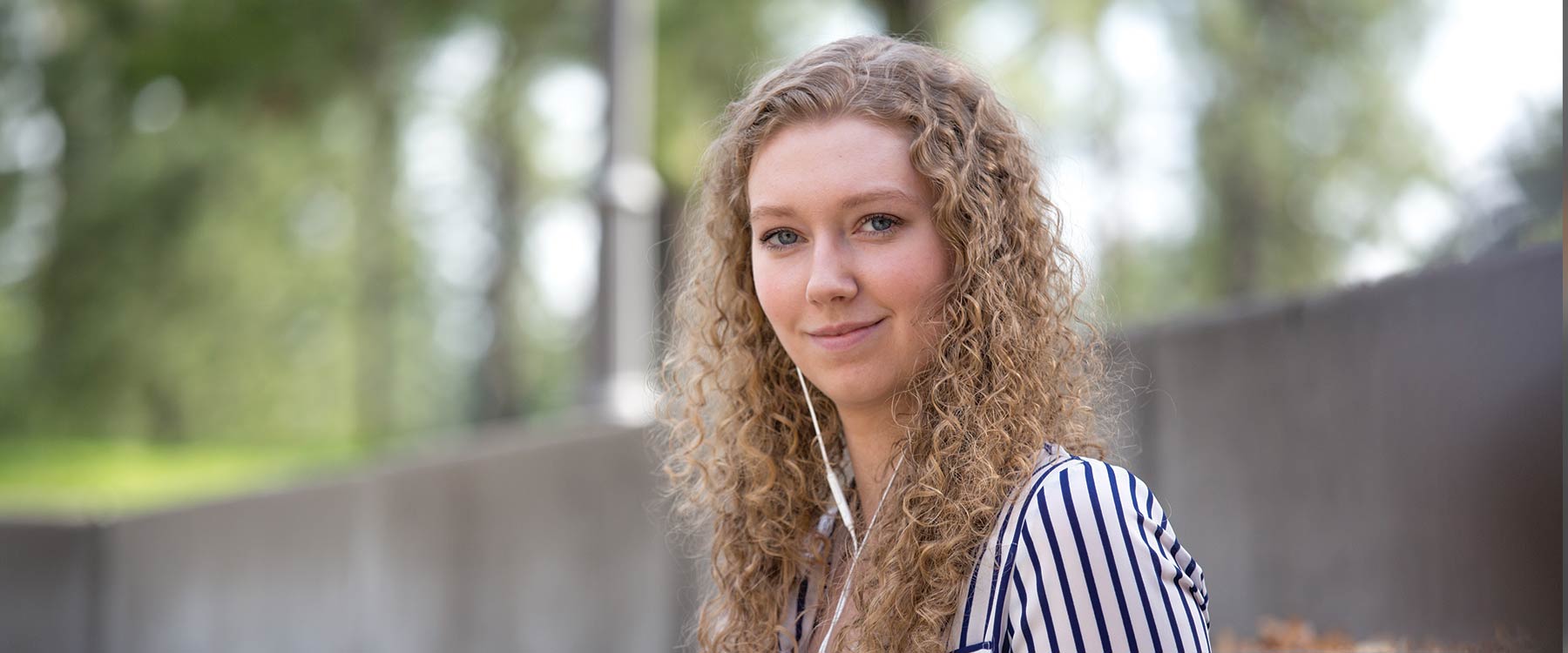 Victoria sits on concrete steps somewhere on campus. She is wearing headphones and smiling at the camera.