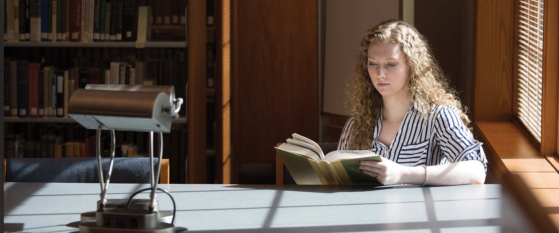 Victoria sits at a table in the library near a window. She stares down at the pages of a book she is reading.