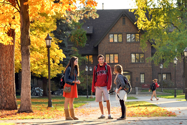 Three students stand outside a stately academic building in fall, talking.