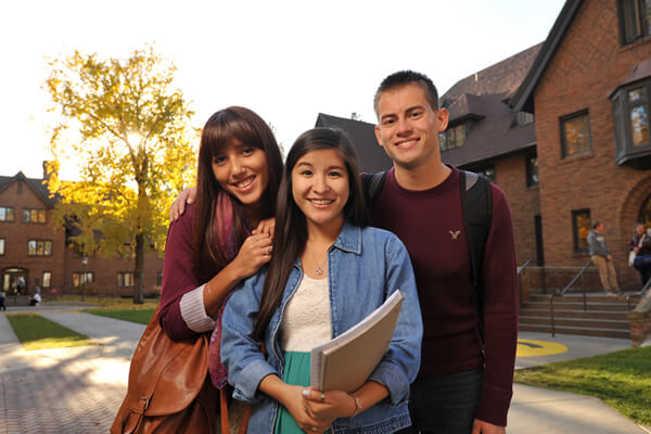 Three students pose and smile together outside of a large brick academic building on a clear day.