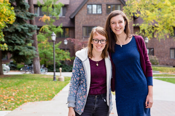 Two students pose side-by-side and smile outside a stately brick academic building on a fall day.