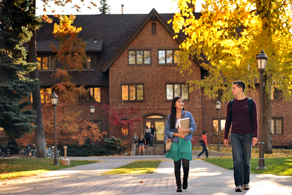 Two students walk together on a tree-lined campus path in autumn, with a brick building in the background.