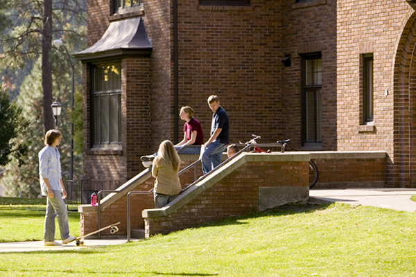 A group of four students linger on and near the steps of a brick building with bicycles parked nearby.