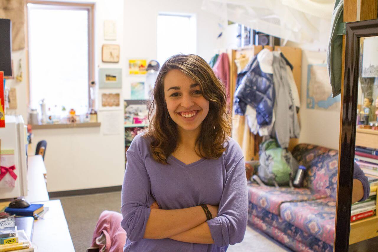 A student stands and smiles in a dorm room with a colorful couch, desk and hung artwork in the background.
