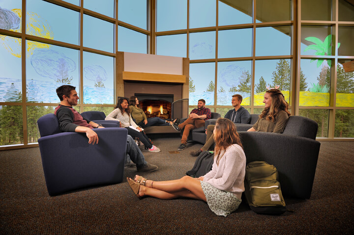 A group of students sit and talk in a cozy room with a fireplace and large windows with a colorful mural.