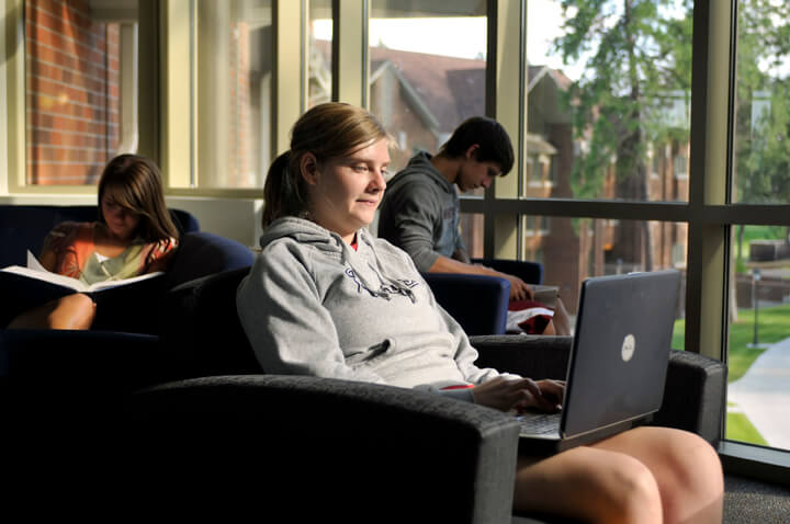 Students study in a sunlit lounge area, with one person using a laptop and two others reading books.