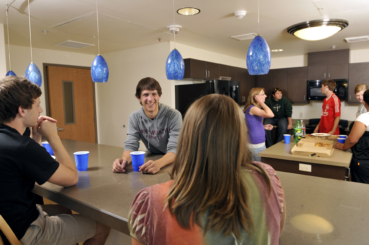 Students gather around two countertops in a large kitchen. A pizza box sits open.
