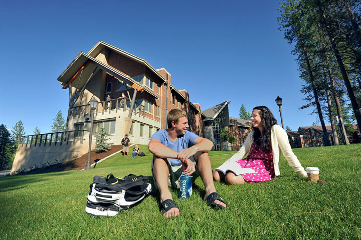 Two students sit on the grass on a hill outside a stately hall with balconies. They look at each other and laugh.