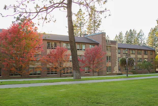Brick campus building behind a row of trees with red and orange fall foliage, viewed from across a grassy lawn.