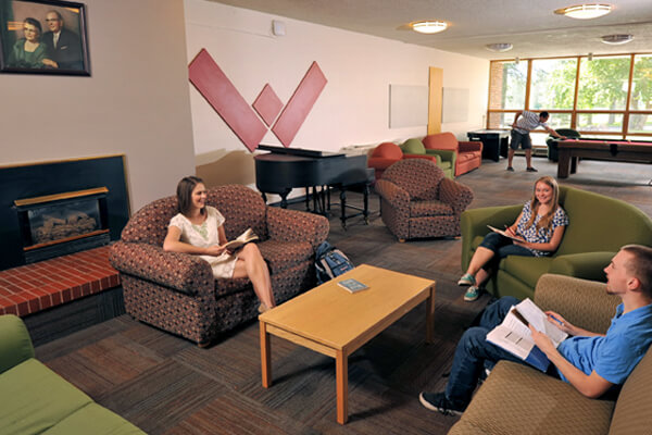 Students sitting on couches and chairs in a lounge area studying and talking, with pool tables and large windows in the background.