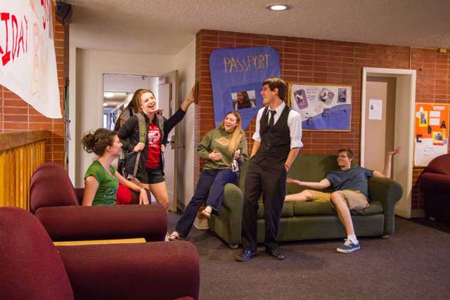 Students gathered in a residence hall lounge, talking near couches and a bulletin board, with a hallway visible in the background.