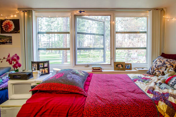Cozy room with a neatly made bed, colorful bedding, string lights and a large windows overlooking trees and a grassy area.