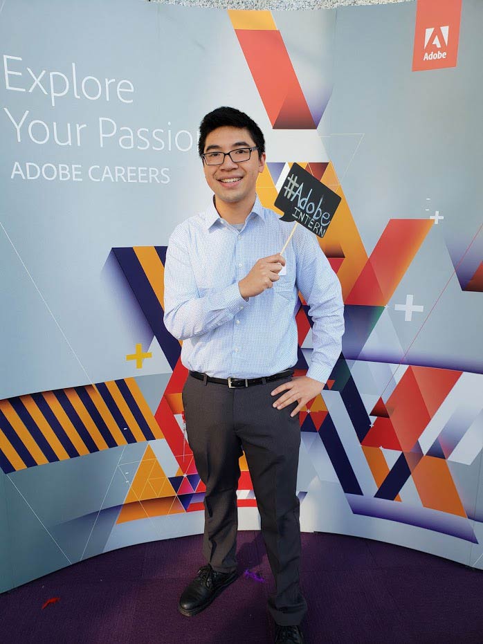 A young adult stands smiling in front of a colorful Adobe Careers backdrop, holding a
