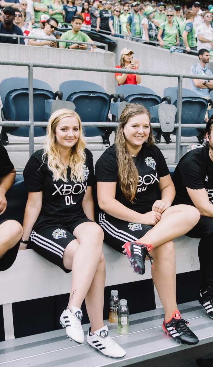 Two young adults wearing soccer jerseys sit on a bench, smiling, with a crowd in the background.