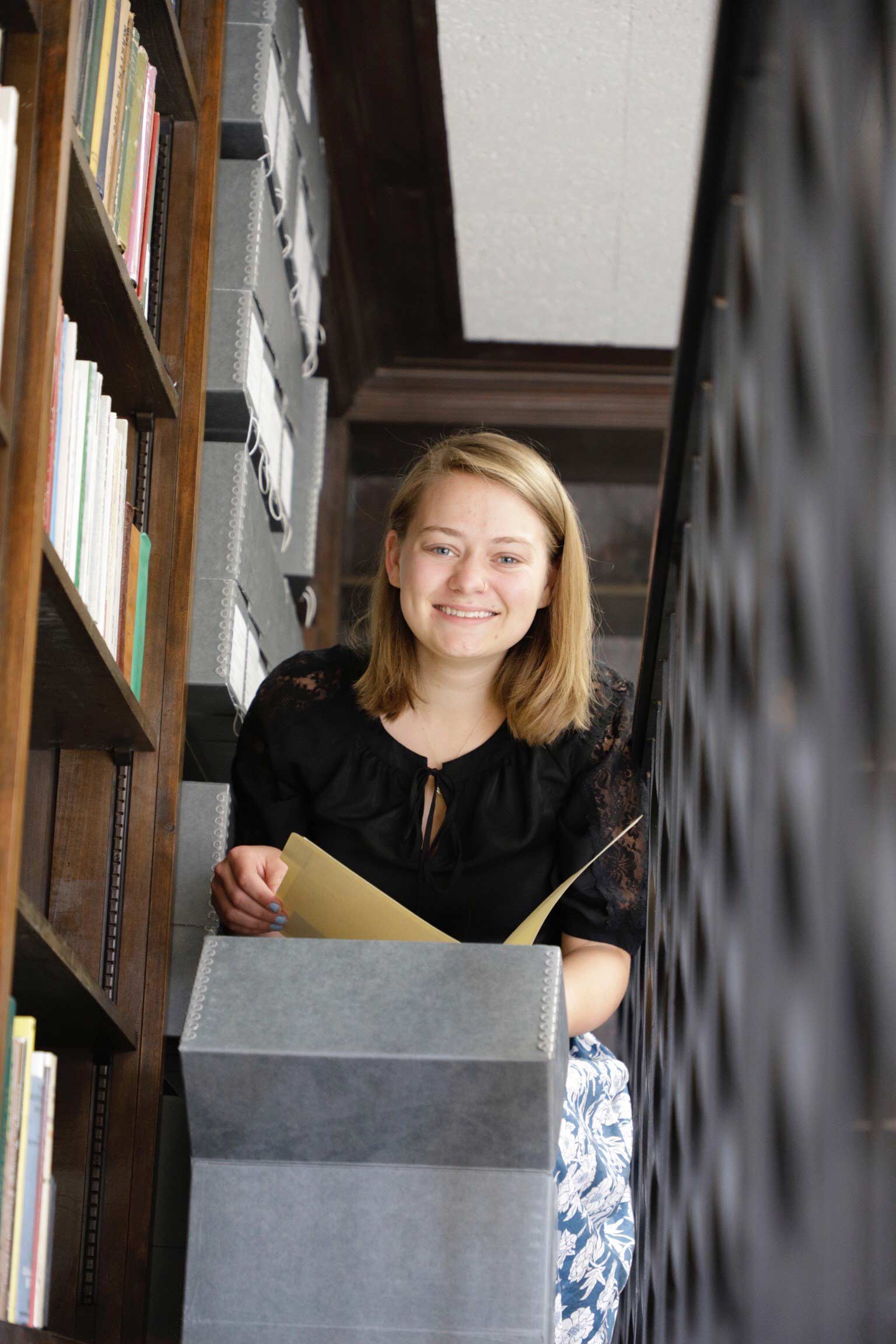 A student smiles and crouches behind a box, holding open a file folder, in a library.