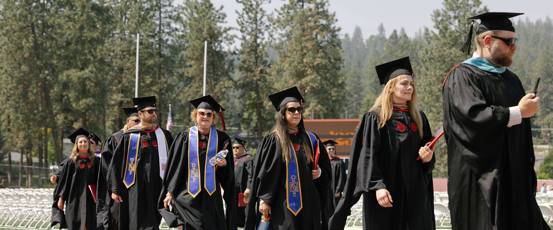 Graduates in caps and gowns walk in procession outdoors on a sunny day, flanked by trees.