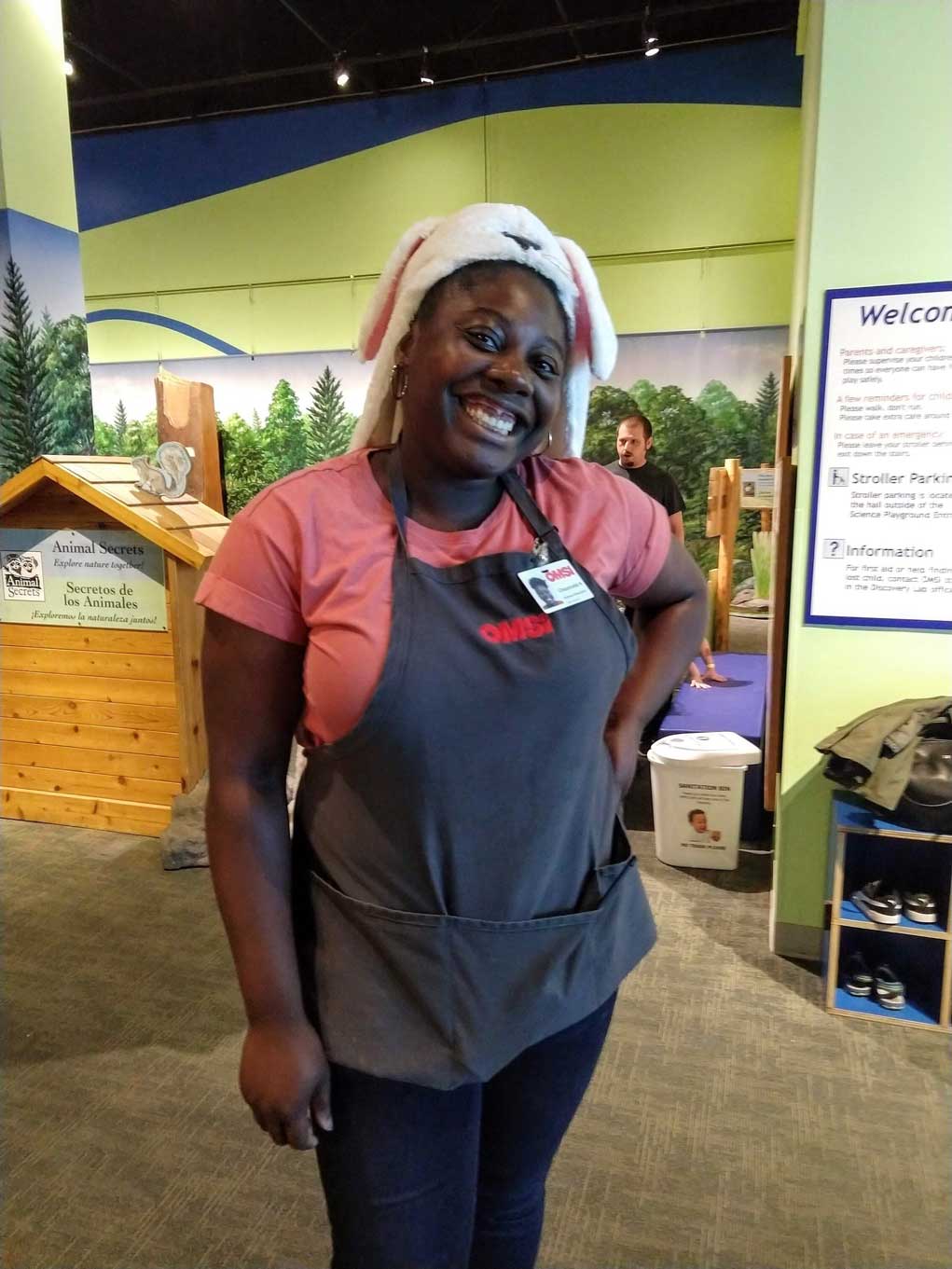 Chauncella Koulibali stands near a museum information booth wearing an apron and bunny hat, smiling.