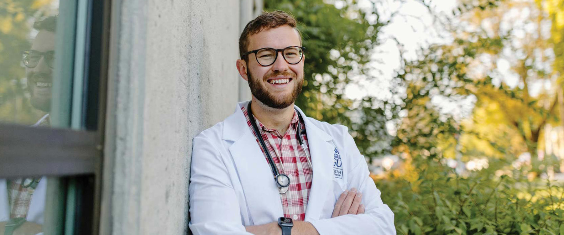 Kevin Glover, wearing a white coat and stethoscope, stands outside, arms crossed, with sunlit trees in the background.