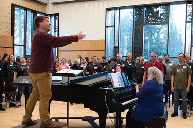 Joshua Chism leads a group of people in a choir practice in a room with large windows.
